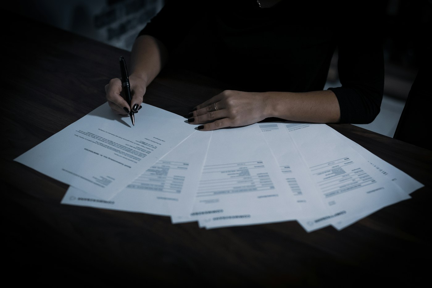 A person signing one of several printed agreements spread across a desk