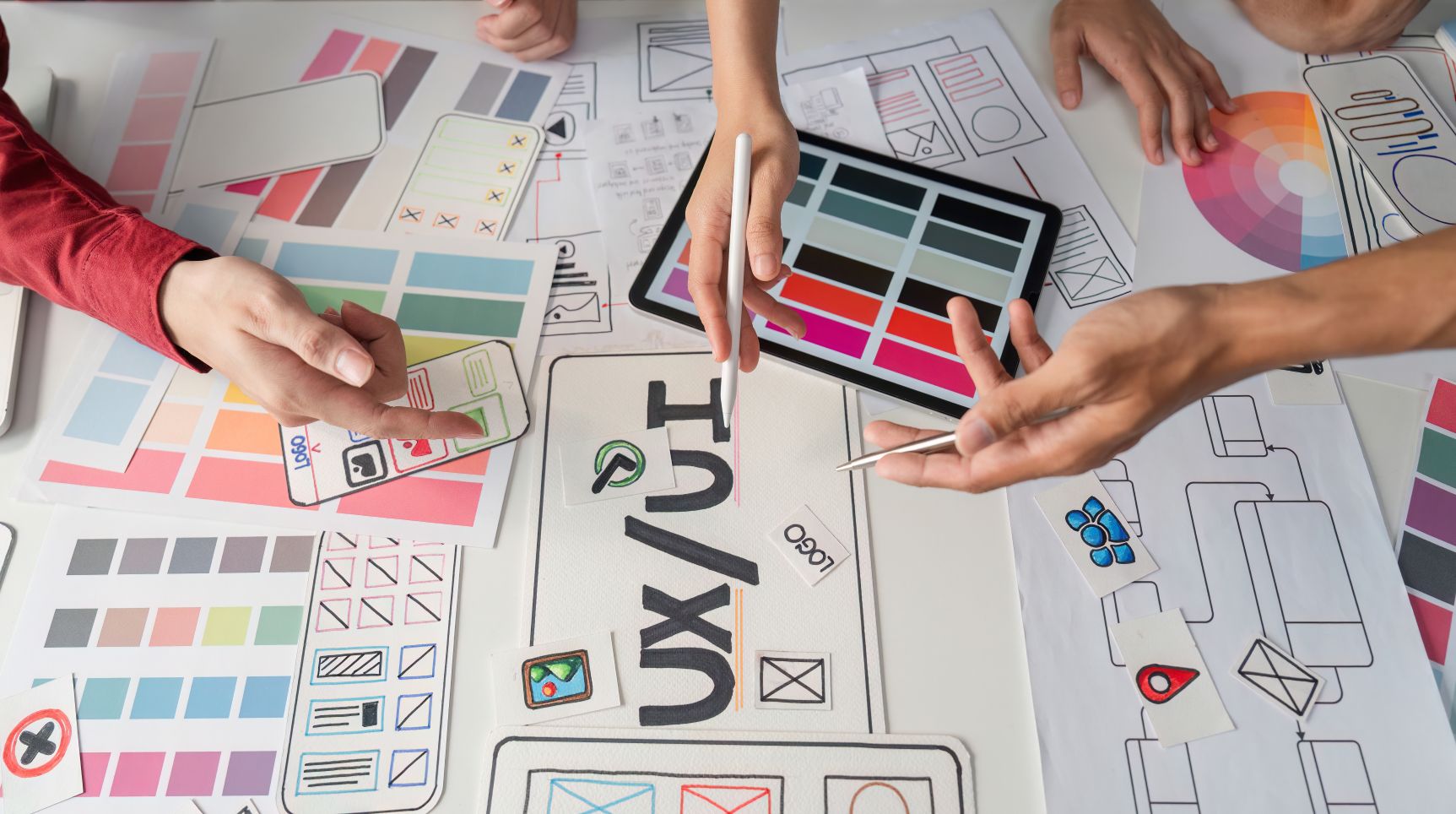 A design team pointing at wireframes, color palettes and a tablet spread around a UX / UI sketch